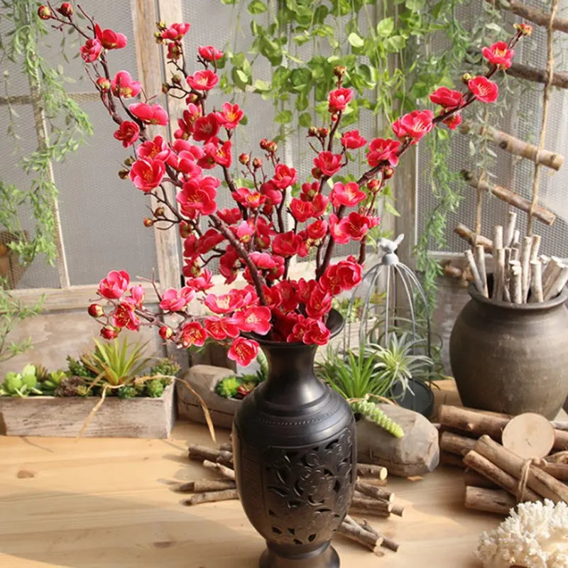 Vibrant red artificial silk cherry blossom branches arranged in a dark vase with greenery and natural wood elements.