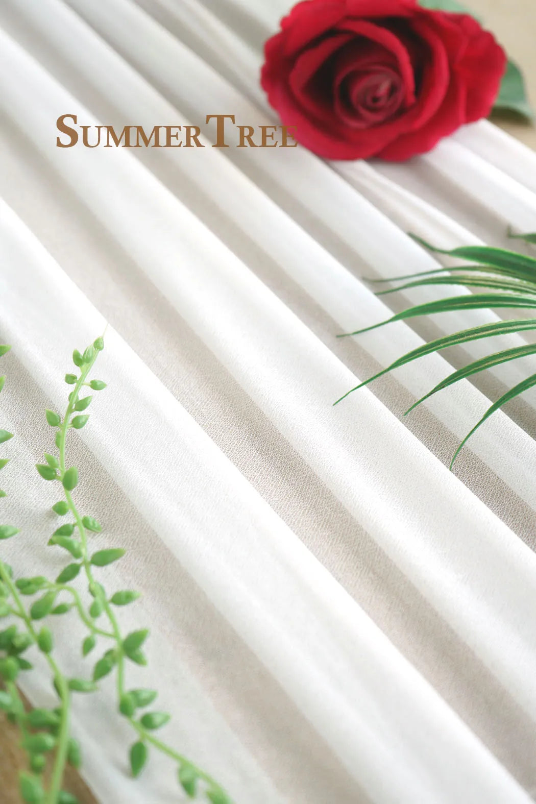 A close-up of elegant cream and white chiffon fabric draped in vertical stripes with a red rose and green foliage.