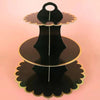 A three-tier black cardboard cupcake stand with gold trim, displayed against a light pink background.