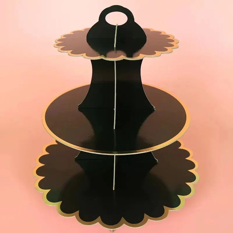 A three-tier black cardboard cupcake stand with gold trim, displayed against a light pink background.