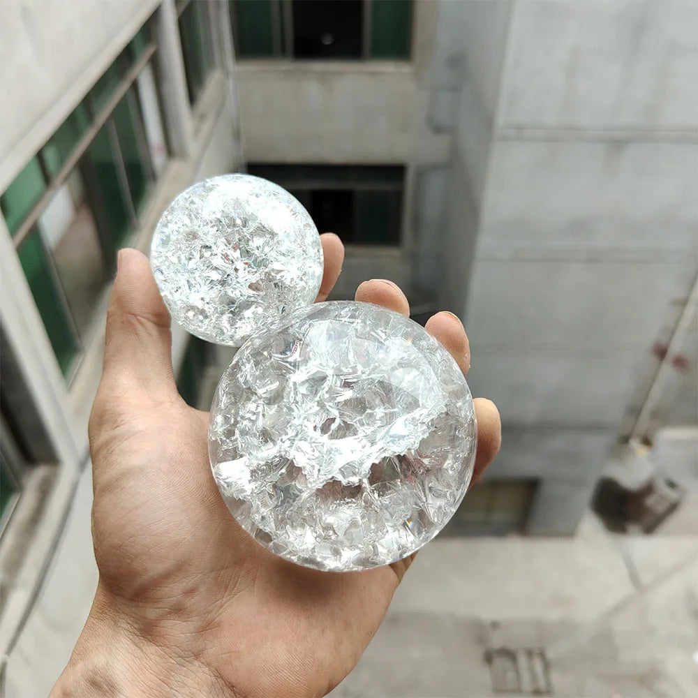 A hand holds a clear, spherical crystal ball water feature ornament against a blurred building background.