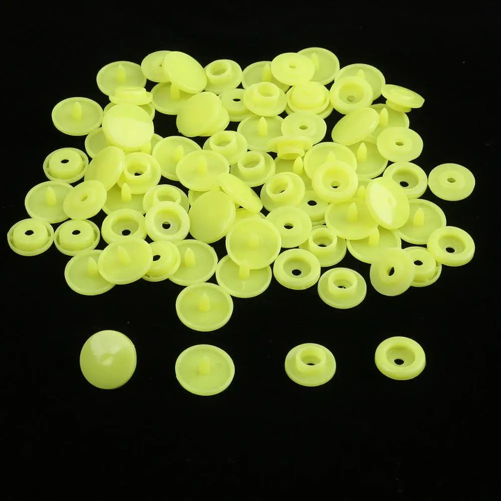 A pile of assorted light yellow snap buttons, measuring 12mm and 10.5mm, scattered on a black surface.