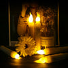 A collection of glowing white LED taper candles in a glass jar and several unlit candles on a dark surface.