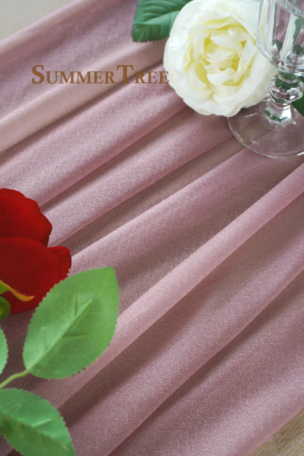 A close-up of pink chiffon fabric draped in vertical folds, with a white rose and green leaves visible.