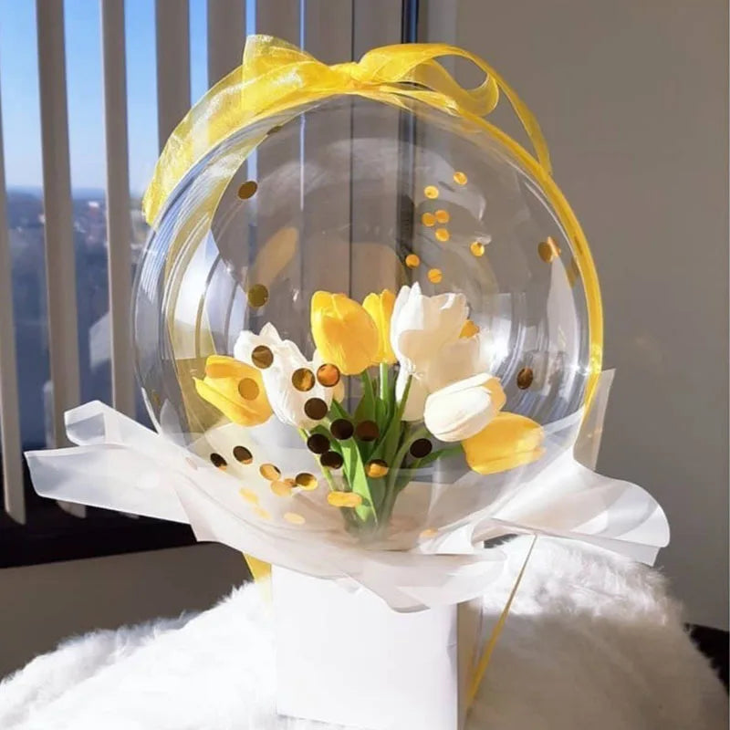 A bouquet of yellow and white tulips is displayed inside a clear balloon tied with a yellow ribbon.