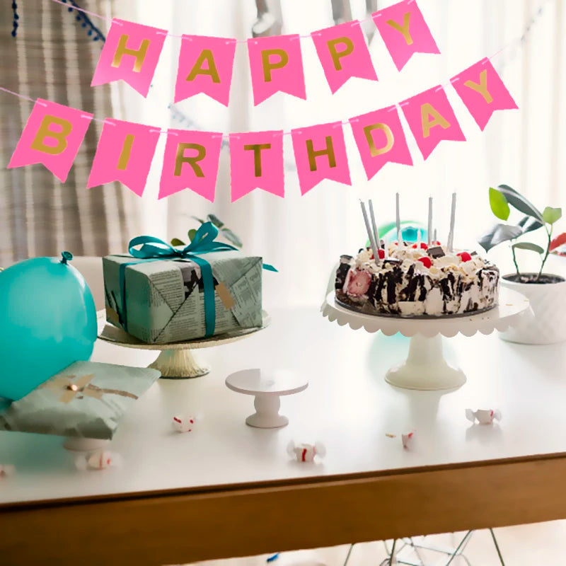 Una pancarta de cumpleaños dorada con banderines rosas cuelga sobre una mesa con un pastel y regalos envueltos.