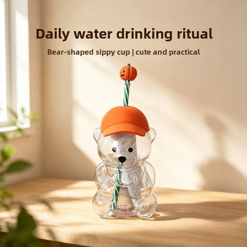 A bear-shaped glass cup with an orange lid and green straw is shown on a wooden surface.