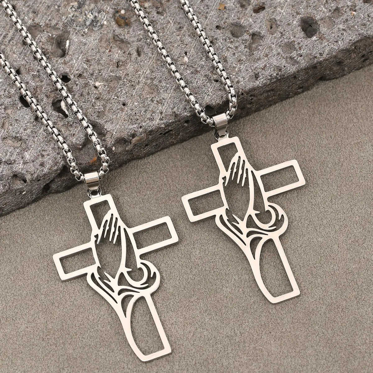 Silver stainless steel cross pendant featuring praying hands design resting on a gray stone surface