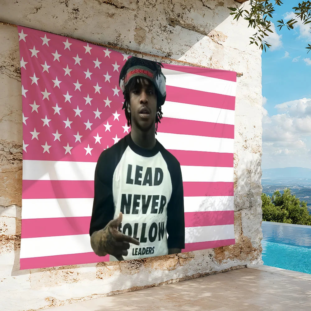 A person wearing a black hoodie and a hat stands in front of a pink and white striped American flag banner on an outdoor wall