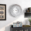 A modern silver wall clock with a textured, sunburst design hangs on a white wall.