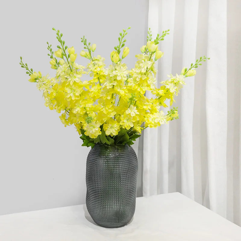 Un bouquet de fleurs artificielles de delphinium jaune vif dans un vase gris texturé sur une surface blanche.