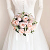 A bridal bouquet of soft pink and blush roses with white accents held by a model in a white dress.