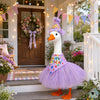 A white duck costume with a purple tutu and a birthday bow standing on a wooden porch.