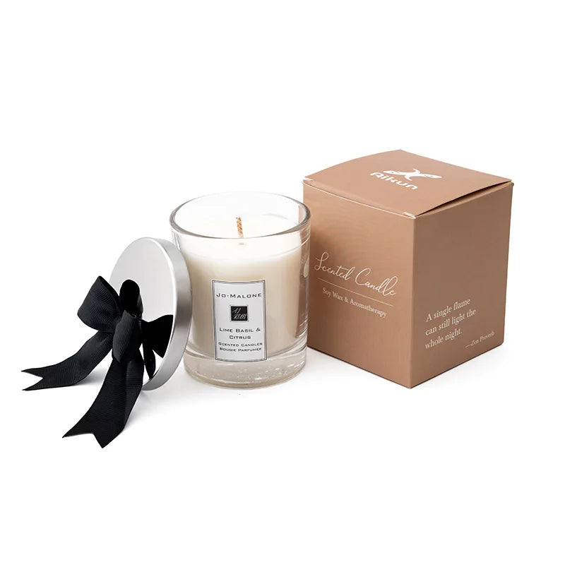 A scented candle in a glass cup next to its kraft paper box with a black ribbon.