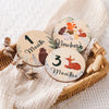 A set of wooden baby milestone cards showing 1 month, newborn, and 3 months, displayed on a cream linen fabric.