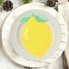 A yellow lemon with a green leaf is centered on a white plate, set on a cream-colored linen napkin.
