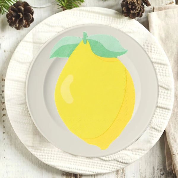 A yellow lemon with a green leaf is centered on a white plate, set on a cream-colored linen napkin.