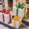 Three white gift boxes wrapped in red, green, and gold ribbons with sparkling lights on a snowy surface