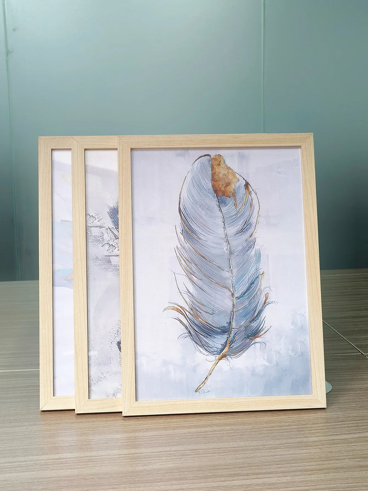 A set of three light wood picture frames with acrylic fronts displaying a watercolor feather painting.