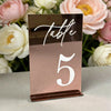 Elegant gold plated mirrored acrylic table number displaying the number 5 on a light surface surrounded by pink and white flo