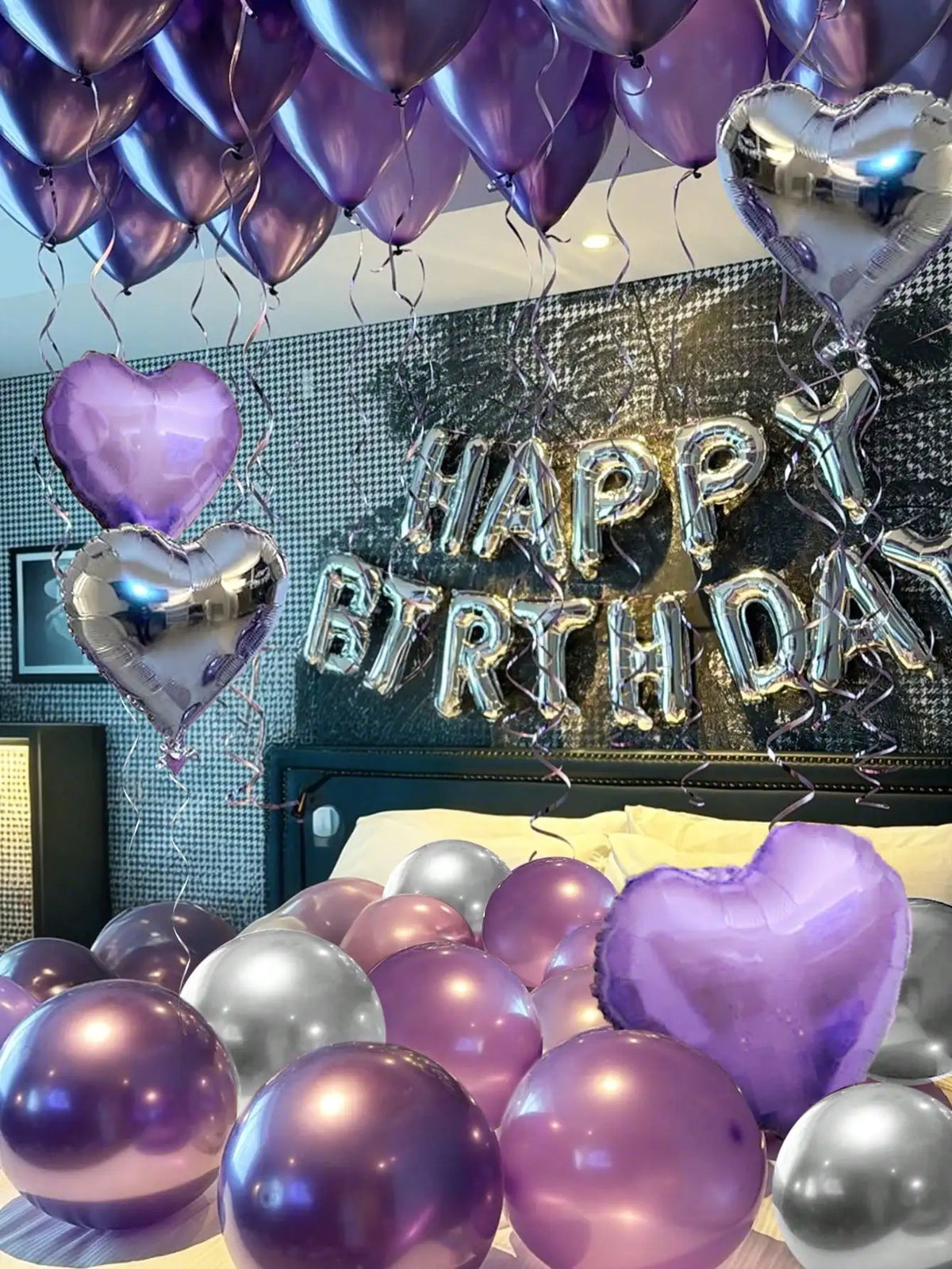 A display of purple and silver metallic balloons, including heart-shaped balloons, decorating a wall for a birthday celebrati