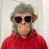 A realistic chimpanzee latex mask with gray fur and brown skin is worn by a person in a red hoodie.