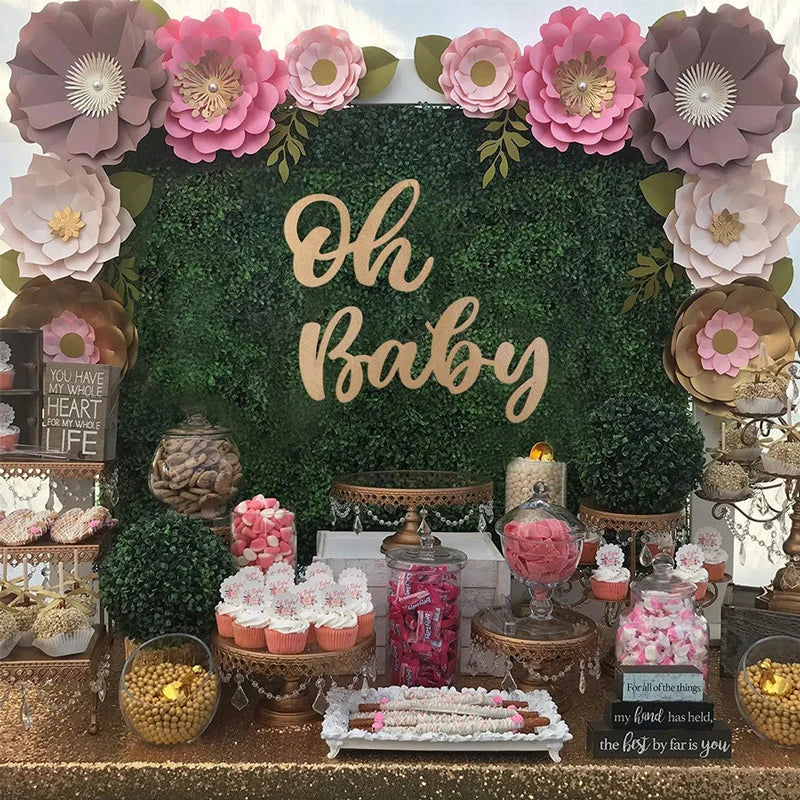 A wooden sign with gold lettering that reads Oh Baby set against a backdrop of green foliage and pink flowers.