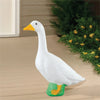 A white goose garden statue with an orange beak and green feet is displayed on a wooden surface next to green foliage.