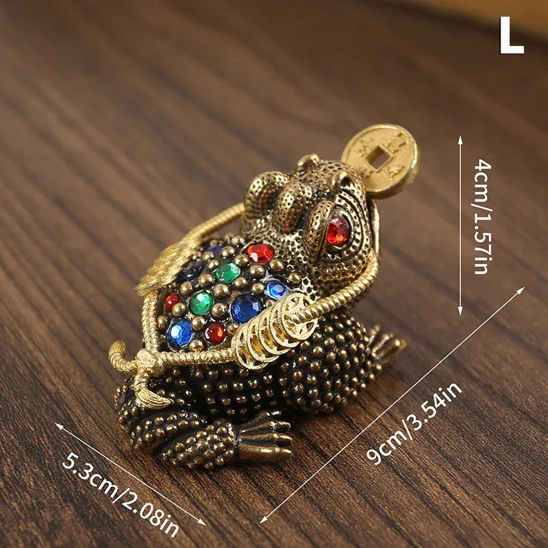 Una pequeña figura de sapo chino de latón con boca dorada y joyas decorativas sobre una superficie de madera oscura.