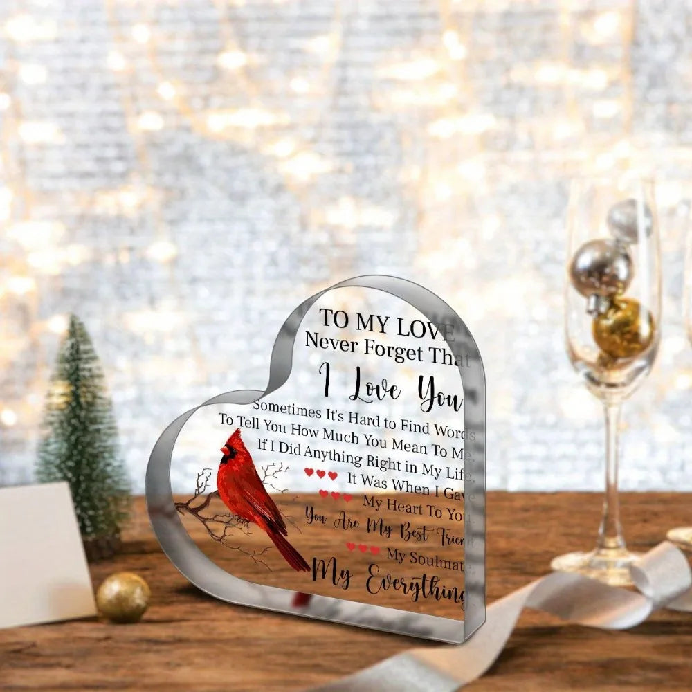 A heart-shaped acrylic plaque with a red bird design and a love message resting on a wooden surface.