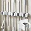 A set of three self-adhesive wall holders for mops, brooms, and long-handled tools mounted on a light-colored wall.