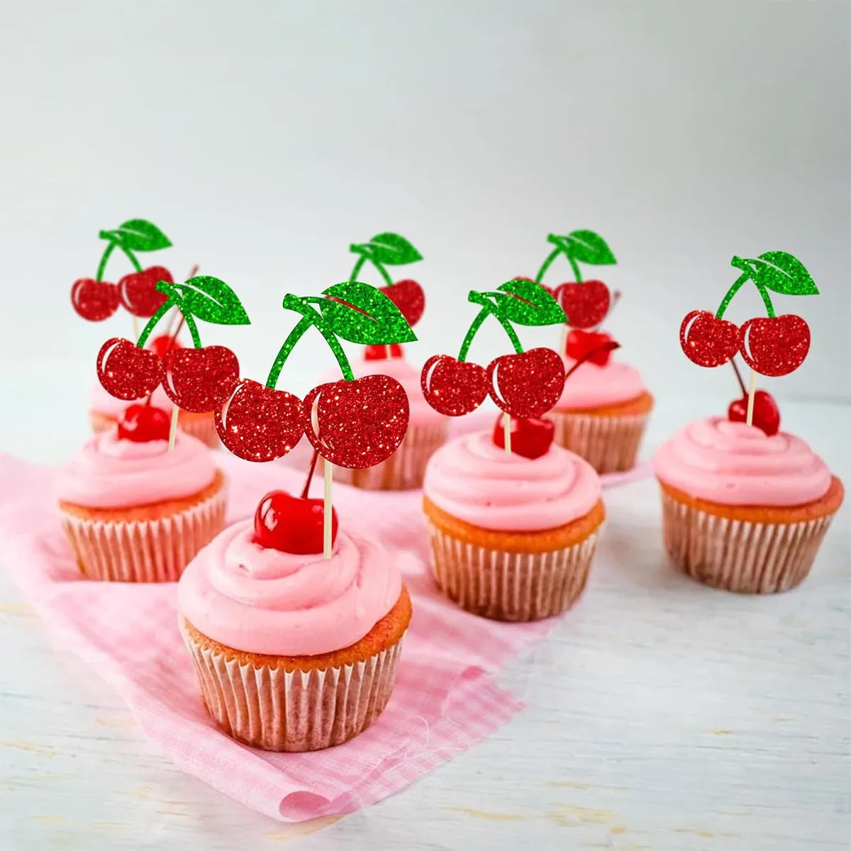 A collection of pink frosted cupcakes decorated with red cherries and green leaves.