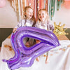 Two young girls smiling while holding a large purple foil number balloon shaped like the number 7.