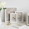 A natural wood picture frame with plexiglass displaying a pink floral arrangement on a white surface.