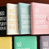 A collection of pink, mint green, beige, and black 100 envelope challenge binders for cash savings organization.