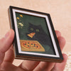 A framed picture featuring a black cat looking at a slice of pizza against a dark green background.