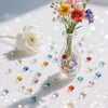 A glass vase holds a bouquet of flowers and scattered colorful acrylic diamond gemstones on a white surface.