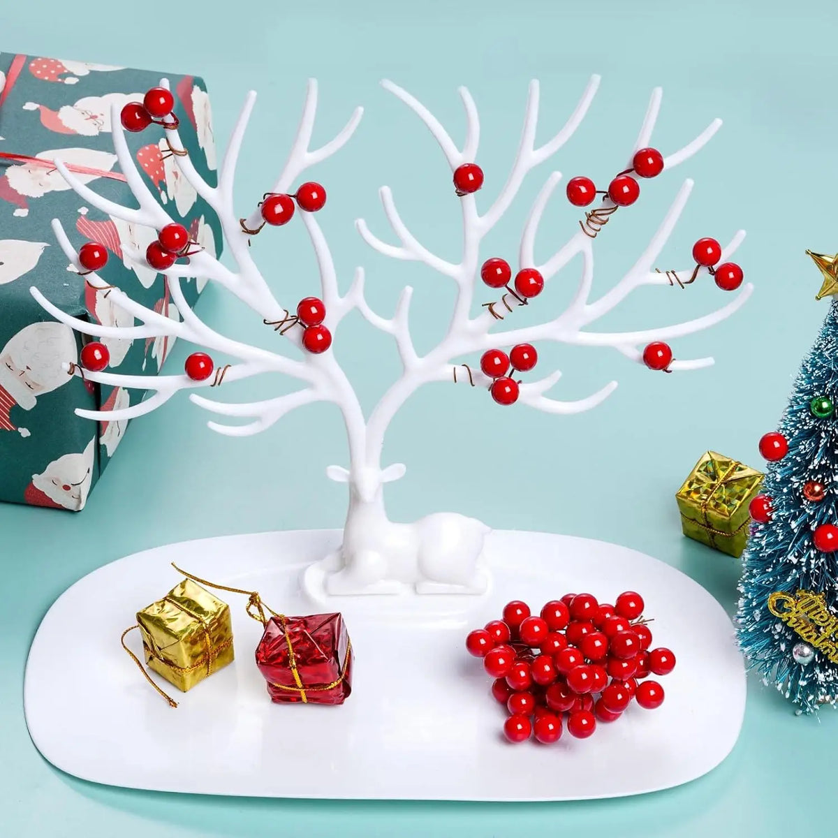 Una rama de árbol artificial blanca decorada con bayas de hieto rojo, mostrada en un plato blanco junto a una caja de regalo 