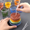 Hands are pouring colored sand pigments into glass containers for a handmade candle wax kit.