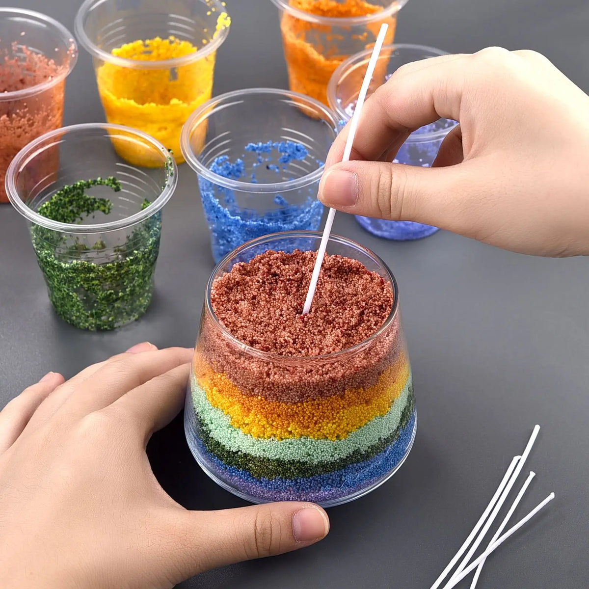 Hands are pouring colored sand pigments into glass containers for a handmade candle wax kit.
