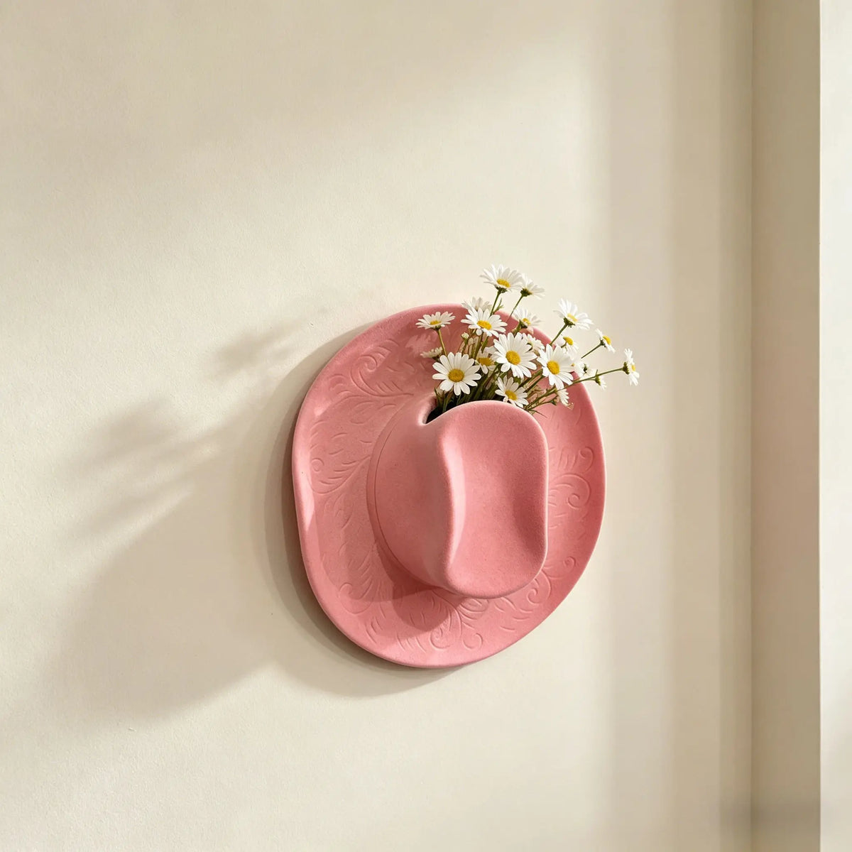 A pink ceramic cowboy hat shaped wall vase with white daisies mounted on a light beige wall.