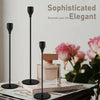 Three black metal taper candle holders displayed on a book next to a perfume bottle and pink roses.