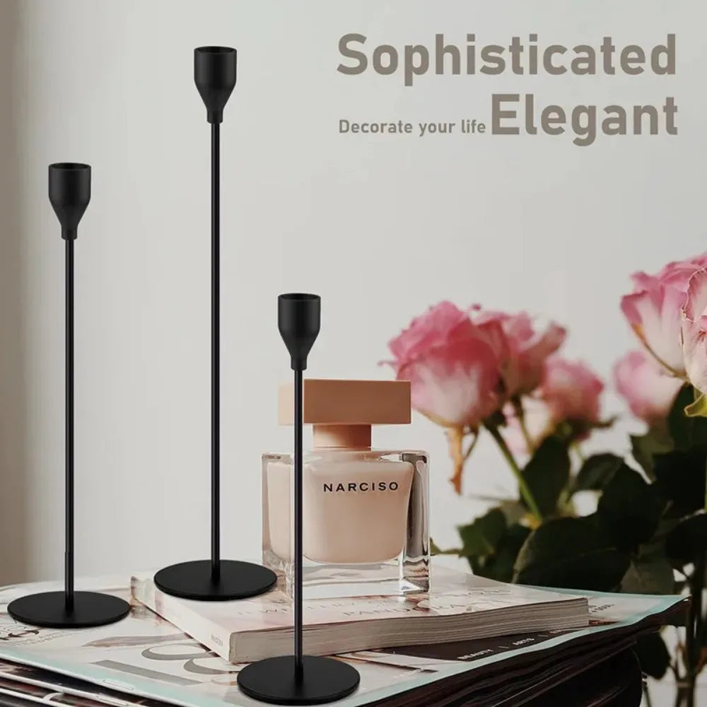 Three black metal taper candle holders displayed on a book next to a perfume bottle and pink roses.