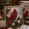 A framed Christmas 3D fabric poke art kit featuring a red bird in a winter forest scene with greenery and snow.