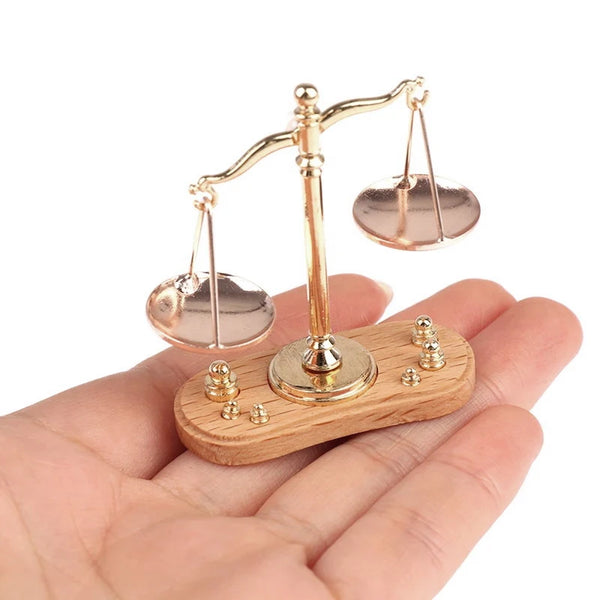 A vintage miniature balance scale with gold-toned arms and plates rests on a wooden base, held in a person's hand.
