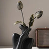 A black ceramic vase holds a dried, closed flower bud against a neutral background.