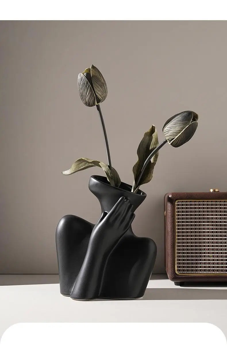 A black ceramic vase holds a dried, closed flower bud against a neutral background.