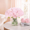 A bouquet of pink silk peony artificial flowers in a clear glass vase on a white table.