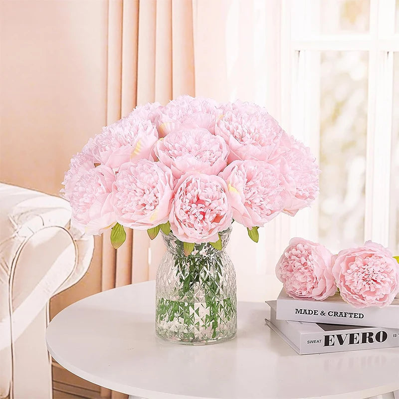 A bouquet of pink silk peony artificial flowers in a clear glass vase on a white table.