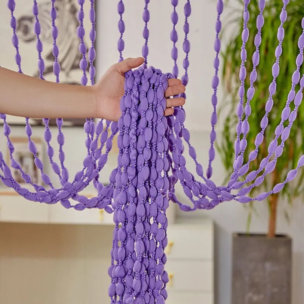 A hand is pulling a purple beaded string curtain with tassels, showcasing the bohemian drape texture.